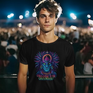Op de afbeelding: Zwart t-shirt met een levendige afbeelding van een gestileerd gezicht met een kroon en de tekst "DECODE THE UNIVERSE" in een neonkleurverloop. Het ontwerp heeft tinten blauw, paars en groen. De persoon staat voor een menigte bij een concert.