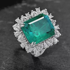 May include: A silver-toned ring featuring a large, rectangular, emerald-green center stone held by four prongs. The center stone is surrounded by a halo of numerous small, pear-shaped, clear white stones set in a metallic frame.