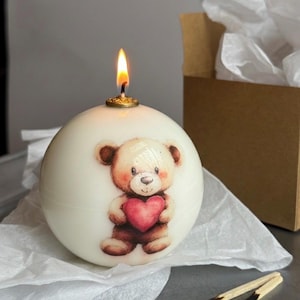 May include: A round, ivory-colored candle with a lit wick. The candle features a painted illustration of a teddy bear holding a red heart. The candle sits on white tissue paper, with a brown box and burnt matches in the background.