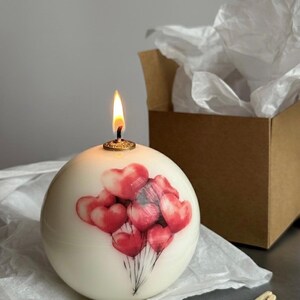May include: A lit, round, ivory-colored candle with a cluster of red heart-shaped balloons printed on it. The candle has a gold-colored wick holder and is sitting on white tissue paper. A brown box and burnt matches are in the background.