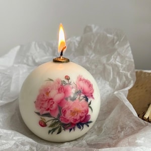 May include: A round, ivory-colored candle with a floral peony design. The candle is lit, with a golden wick holder and a bright flame. The candle rests on white tissue paper, suggesting a decorative or gift item.