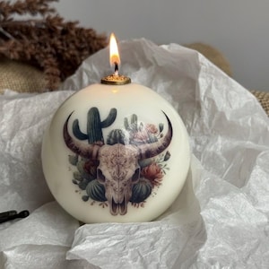 May include: A round, ivory-colored candle with a lit wick. The candle features a detailed illustration of a bull skull, cacti, and flowers. The design is in shades of brown, green, and red. The candle sits on white tissue paper.