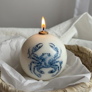May include: A spherical, white candle with a blue crab design. The candle is lit, with a flame at the top. It sits in a woven basket lined with white paper. The candle is a decorative item.