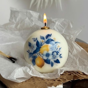 May include: A spherical candle with a lit wick, featuring a lemon and floral design in blue and yellow. The candle sits on white tissue paper and brown cardboard. Two used matches are visible.