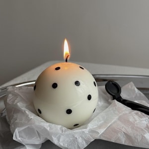 May include: A spherical white candle with black polka dots is lit with a flame. The candle sits on a silver tray with a piece of white tissue paper. A black candle snuffer and two burnt matches are also on the tray.