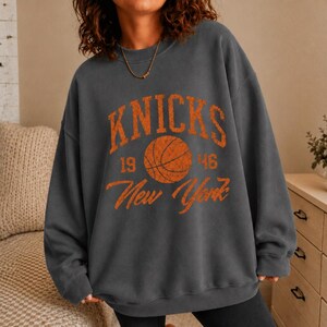 May include: A charcoal gray crewneck sweatshirt featuring a distressed orange graphic print on the chest. The design includes the text "KNICKS" above a basketball illustration, with "19" and "46" on either side and "New York" written below.