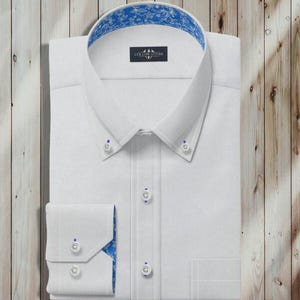 Könnte beinhalten: Gefaltetes weißes Hemd mit Button-Down-Kragen und blau gemustertem Futter an Kragen und Manschetten. Das Hemd hat blaue Knöpfe und eine kleine Tasche. Das Markenlogo ist sichtbar.