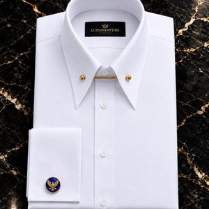 Può includere: Una camicia elegante bianca con colletto a punta, una barretta da colletto dorata, un'abbottonatura frontale e un polsino doppio chiuso da un gemello rotondo blu e oro con un emblema a forma di aquila. L'etichetta interna riporta la scritta LUXLINEATTIRE.