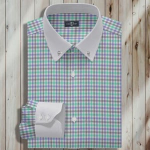 May include: A button-down shirt with a white collar and cuffs. The shirt features a checkered pattern in shades of green, purple, and gray. The shirt is neatly folded on a wooden surface.