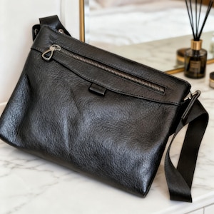 Puede incluir: Bolso bandolera de cuero negro con superficie texturizada. Cuenta con cierre de cremallera plateada y correa negra ajustable. El bolso rectangular presenta un diseño minimalista.