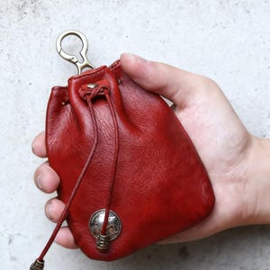 Puede incluir: Una pequeña bolsa de cuero roja con cierre de cordón y cierre metálico. La bolsa presenta un emblema metálico decorativo y se sostiene en una mano. La bolsa está diseñada para guardar objetos pequeños.