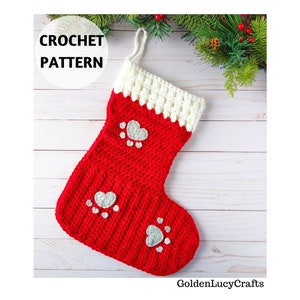 May include: Red and white crocheted Christmas stocking with a silver heart and paw print design. The stocking is hanging on a white wooden background with green pine branches.