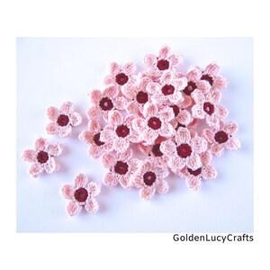 May include: A collection of small, five-petaled crochet flowers in light pink with dark maroon centres. The flowers are arranged in a cluster with some scattered individually. The text GoldenLucyCrafts is visible in the bottom right corner.