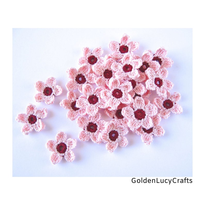 May include: A collection of small, five-petaled crochet flowers in light pink with dark maroon centres. The flowers are arranged in a cluster with some scattered individually. The text GoldenLucyCrafts is visible in the bottom right corner.