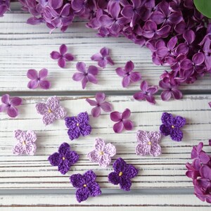May include: A collection of small, four-petaled crochet flowers in shades of light and dark purple, each featuring a tiny yellow centre, arranged alongside fresh, natural purple lilac blossoms on a white, distressed wooden surface.