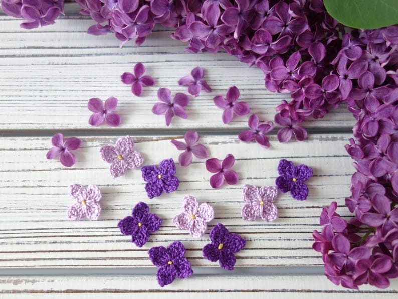 May include: A collection of small, four-petaled crochet flowers in shades of light and dark purple, each featuring a tiny yellow centre, arranged alongside fresh, natural purple lilac blossoms on a white, distressed wooden surface.