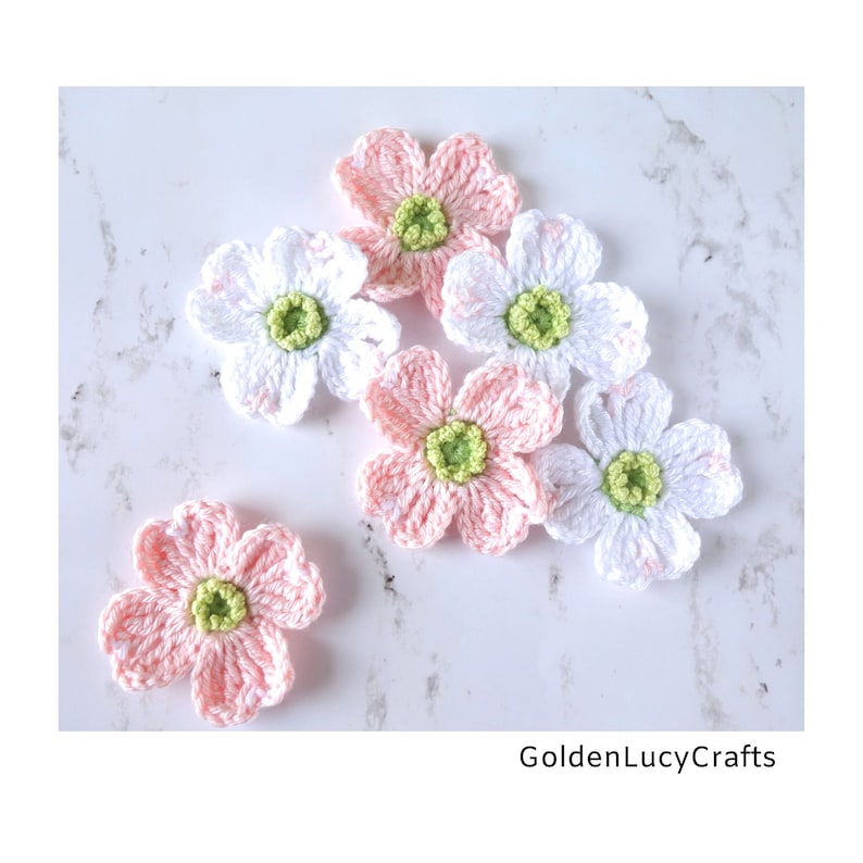 May include: Six handmade crochet flowers in shades of white and light pink, each featuring four rounded petals and a textured lime green centre. The text GoldenLucyCrafts is printed in black in the bottom right corner of the image.