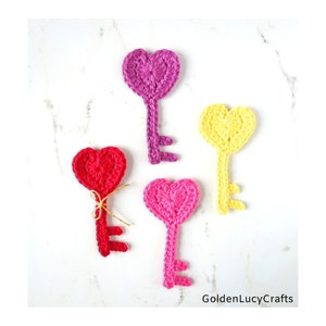 May include: Four crocheted heart-shaped keys in different colors: pink, red, yellow, and purple. The keys are arranged on a white marble surface. The text "GoldenLucyCrafts" is visible in the bottom right corner of the image.