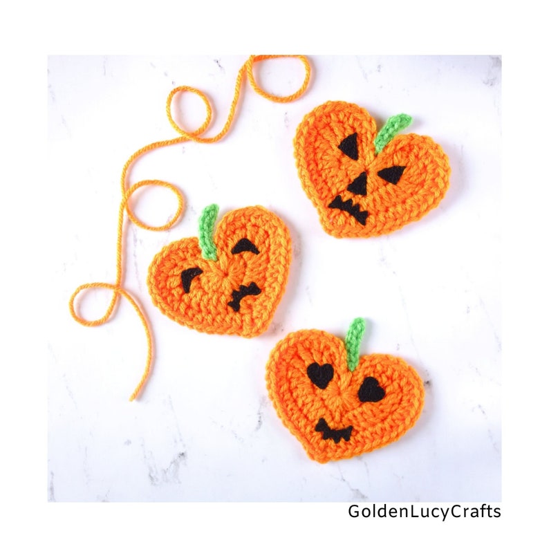 May include: Three crocheted orange heart-shaped pumpkins with black eyes and mouths. The pumpkins have green stems. The pumpkins are on a white surface.