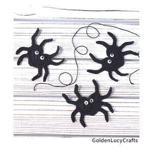 May include: Three black crocheted spiders with white googly eyes on a white wooden background. The spiders are connected by a black string.