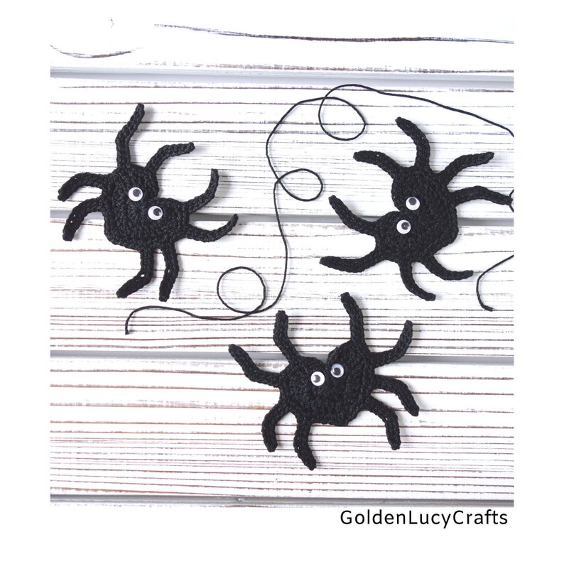 May include: Three black crocheted spiders with white googly eyes on a white wooden background. The spiders are connected by a black string.