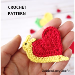 May include: A handmade crochet snail applique with a red heart-shaped shell and yellow body. The text "CROCHET PATTERN" is visible in the image. The snail is held in a hand, with other crochet hearts in the background.