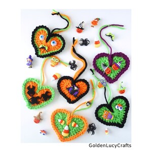 May include: Five crocheted Halloween-themed hearts with various embellishments, including googly eyes, spiders, bats, and candy corn. The hearts are in shades of green, orange, purple, and black. The text "GoldenLucyCrafts" is visible at the bottom of the image.