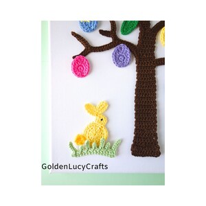 Puede incluir: Un &aacute;rbol de Pascua tejido a crochet con ramas marrones y huevos de colores. Un conejito tejido a crochet amarillo se sienta en la hierba verde en la base del &aacute;rbol. El texto "GoldenLucyCrafts" es visible en la parte inferior de la imagen.