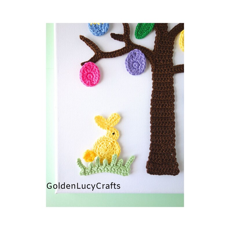 Puede incluir: Un &aacute;rbol de Pascua tejido a crochet con ramas marrones y huevos de colores. Un conejito tejido a crochet amarillo se sienta en la hierba verde en la base del &aacute;rbol. El texto "GoldenLucyCrafts" es visible en la parte inferior de la imagen.