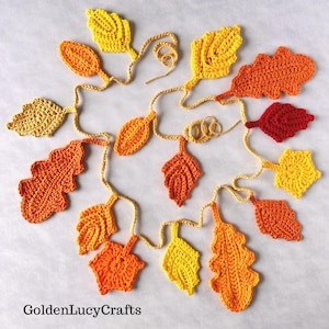 Peut inclure: Une guirlande au crochet composée de diverses feuilles aux couleurs automnales, dans des tons jaune, orange et rouge, reliées par une fine ficelle. Le texte GoldenLucyCrafts est imprimé en noir en bas à gauche de l'image.