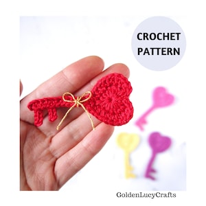 May include: A red crocheted heart-shaped key with a gold ribbon. The key is being held in a hand. The image also shows three other crocheted keys in the background, one pink, one yellow, and one purple.