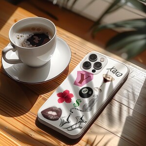May include: A white phone case with various stickers, including a red flower, lips, and the word "xoxo." A cup of coffee sits on a saucer next to the phone case on a wooden surface.