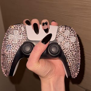 May include: A black and white video game controller featuring two hand grips completely covered in small, light pink, reflective rhinestones. The controller includes standard directional buttons, two black analog sticks, and a central touch-sensitive panel.