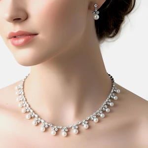 May include: A silver-toned necklace and matching earrings set. The necklace features a delicate design of clear crystals and dangling faux pearls. The earrings have a similar design, with a single pearl drop. The jewelry is designed to be worn for special occasions.