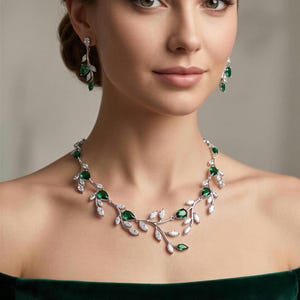 May include: A silver-toned necklace and matching drop earrings featuring a vine-like design. The jewelry set is adorned with marquise and pear-shaped clear white stones and vibrant green teardrop stones arranged in a botanical leaf pattern.