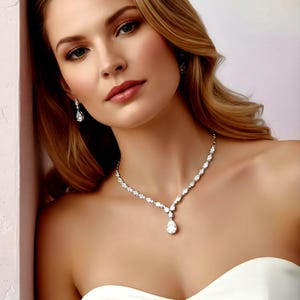Può includere: Una donna indossa una scintillante collana di diamanti e orecchini abbinati. La collana presenta una serie di piccoli diamanti rotondi che conducono a un pendente a forma di goccia più grande. Anche gli orecchini hanno un diamante a forma di goccia. La donna indossa un abito bianco.