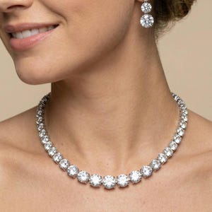 Puede incluir: Un collar brillante y un juego de aretes a juego. El collar presenta una serie de piedras preciosas redondas y claras. Los aretes tienen tres piedras redondas graduadas. La joyería está sobre un fondo neutro.