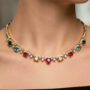 May include: A gold-toned necklace featuring a series of multi-colored gemstones including red, blue, green, and yellow stones in various shapes such as rectangles and a central heart. The metal links are accented with small white stones throughout.