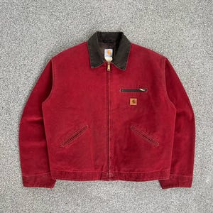 Op de afbeelding: Een rode Carhartt-jas met een bruine kraag en ritssluiting. De jas heeft twee voorzakken en een kleine vierkante logopatch op de borst. De jas is gemaakt van een duurzaam materiaal.