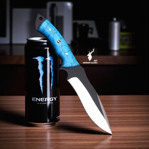 May include: A black and silver knife with a blue handle, resting on a wooden surface next to a black energy drink can. The knife has a curved blade and a handle with a blue, cracked pattern. The energy drink can has the word "ENERGY" in white.