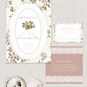 May include: A baby shower invitation set featuring a large card with an apple illustration and the text the apple of our eye, a diaper raffle ticket, and a books for baby card, all decorated with floral patterns and white lace accents near a silver rattle.
