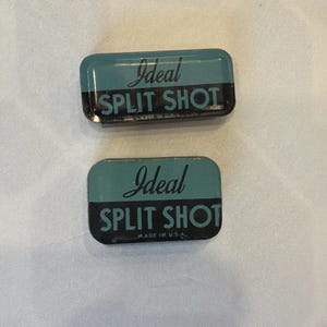 May include: Two rectangular, teal and black metal containers with rounded corners. The text "Ideal SPLIT SHOT" is printed on each container in white script. The bottom container reads "MADE IN U.S.A."