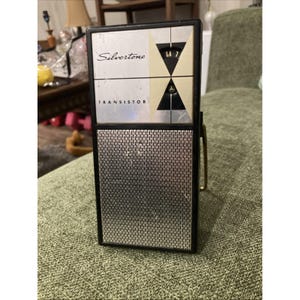 Puede incluir: Radio transistor Silvertone vintage, de color plateado y negro. La radio tiene una rejilla de altavoz plateada, un asa negra y la palabra "TRANSISTOR" impresa en la parte delantera. La cara de la radio tiene la palabra "Silvertone".