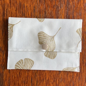 May include: A rectangular fabric pouch featuring an off-white base colour with a repeating pattern of gold ginkgo biloba leaves. The pouch is secured with a single, centred, light beige snap button closure on the front flap of the item.
