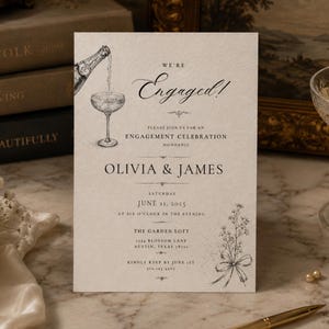 May include: A cream-colored engagement invitation card featuring black ink illustrations of a champagne bottle pouring into a coupe glass and a floral bouquet. The text reads: We're Engaged! Please join us for an engagement celebration honoring Olivia & James.