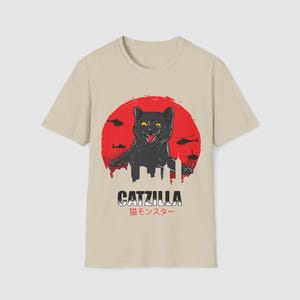 Può includere: T-shirt beige con un'illustrazione di un gatto nero, la scritta "CATZILLA" e caratteri giapponesi. Il gatto è davanti a un sole rosso con elicotteri e una silhouette di città.