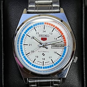 Könnte beinhalten: Eine silberfarbene Seiko 5 Automatik-Armbanduhr mit weißem Zifferblatt. Die Uhr hat blaue und orangefarbene Akzente, eine Tages- und Datumsanzeige und ein Edelstahlarmband. Das Zifferblatt zeigt den Text "SEIKO 5 AUTOMATIC".