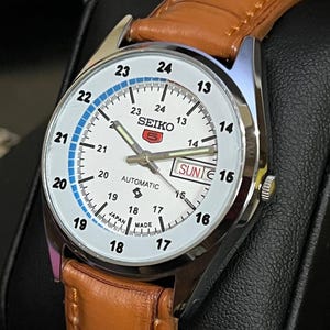 Könnte beinhalten: Eine silberfarbene Seiko 5 Automatik-Uhr mit weißem Zifferblatt, schwarzen Ziffern und Zeigern sowie einer Tages- und Datumsanzeige. Sie hat ein braunes Lederarmband. Auf dem Zifferblatt stehen "SEIKO" und "AUTOMATIC".