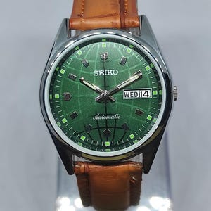 Puede incluir: Reloj de pulsera automático Seiko con esfera verde y correa de cuero marrón. La esfera muestra el día y la fecha, con la palabra "Automatic" impresa debajo de las manecillas. La caja del reloj es de color plateado.