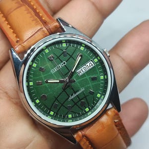 Op de afbeelding: Een Seiko automatisch polshorloge met een groene wijzerplaat en een bruine leren band. De wijzerplaat toont de dag en datum, met het woord "Automatic" onder de wijzers. Het horloge heeft een zilverkleurige kast.